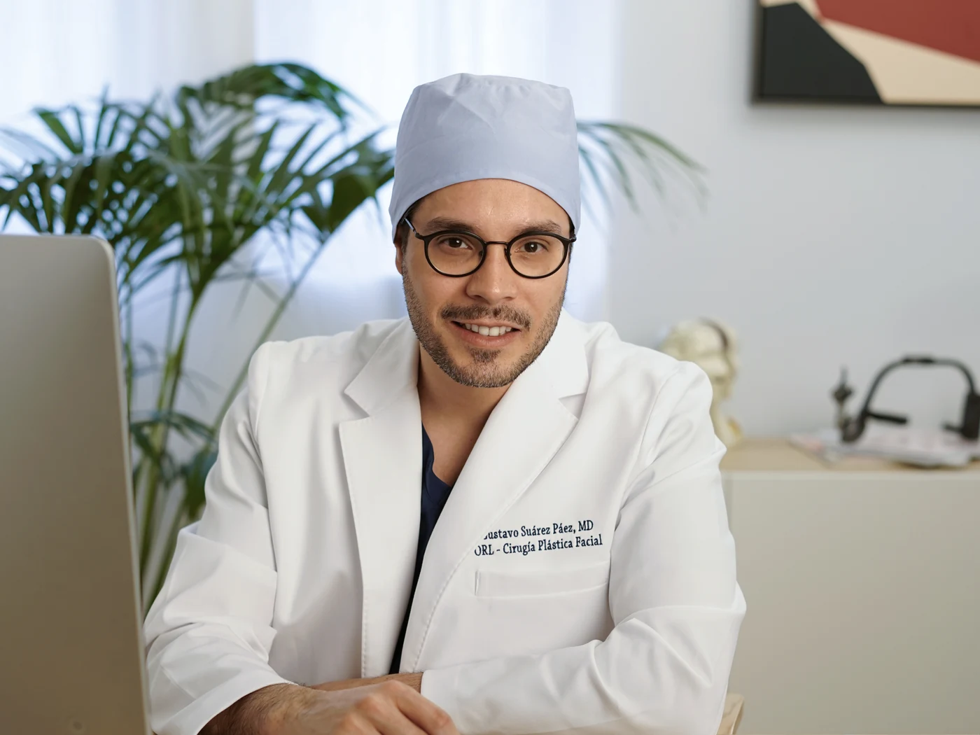 El Dr. Gustavo Suárez, con gorro y bata, sonríe a la cámara sentado ante su mesa de consulta